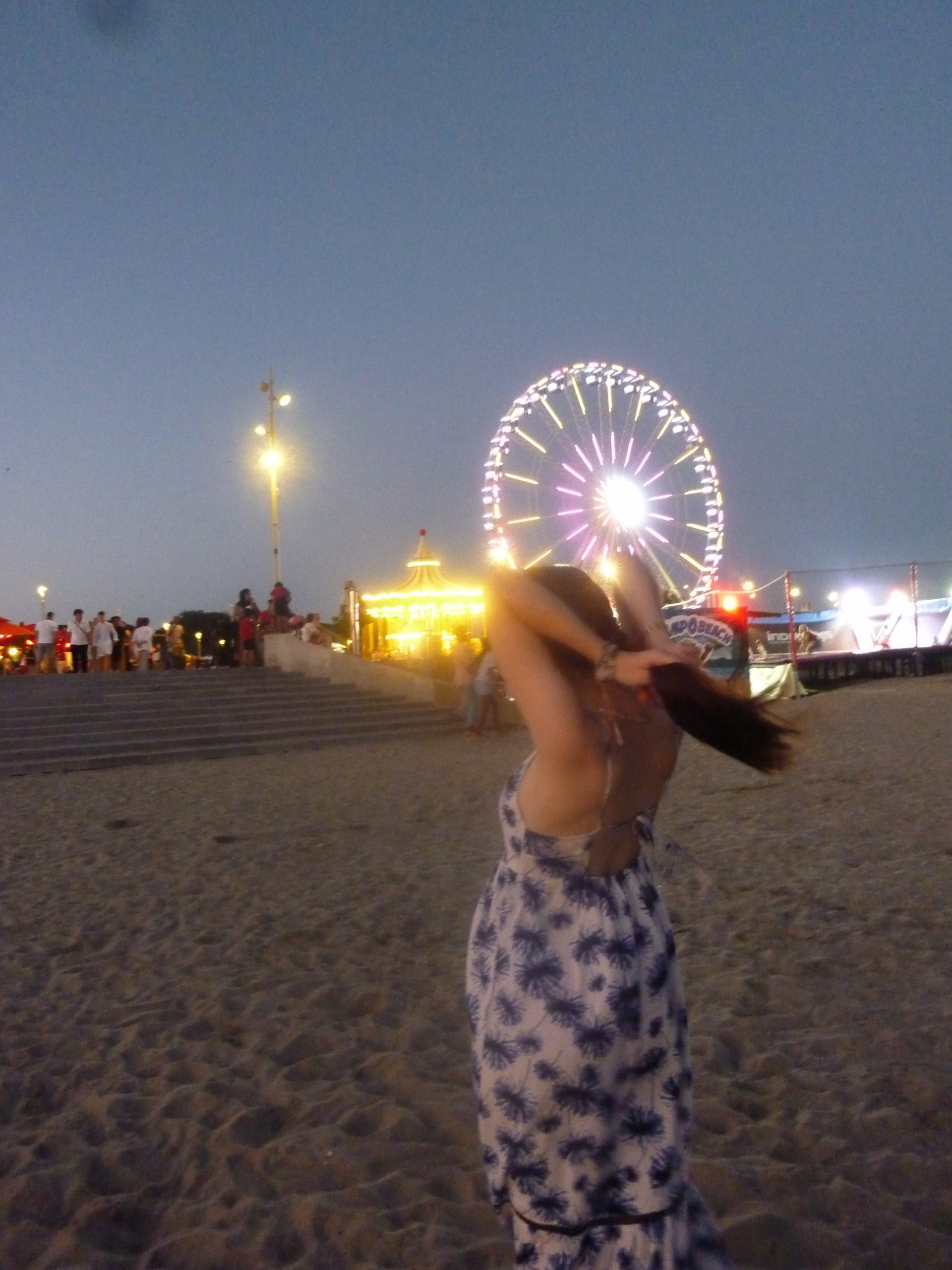 Photo d'une femme les mains dans les cheveux sur la plage de nuit, avec une grande roue et des escaliers en arrière plan
