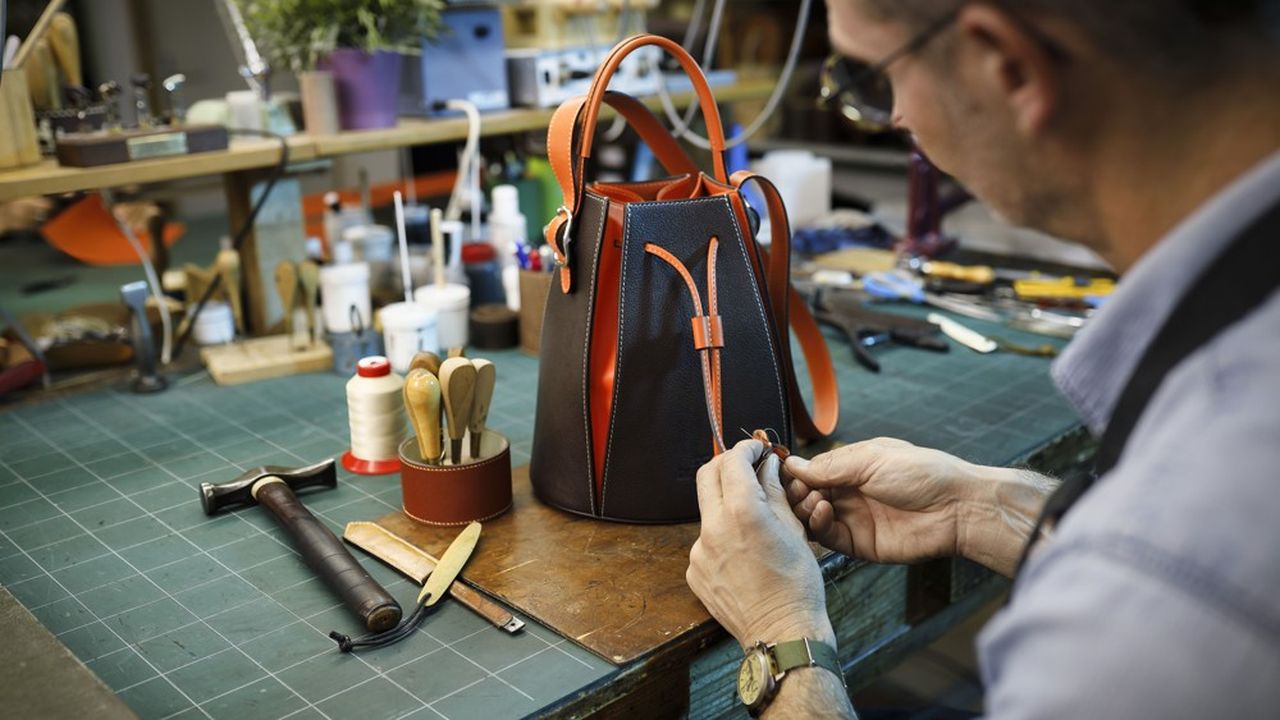 Dans son atelier, un artisan maroquinier travaille avec précision sur les finitions d'un sac seau bicolore (noir et orange). Autour de lui, l'établi regorge d'outils traditionnels comme des marteaux, des alènes et du fil de couture, témoignant d'un savoir-faire artisanal authentique. Cette scène illustre la qualité et la durabilité de la fabrication manuelle, à l'opposé de la production de masse.