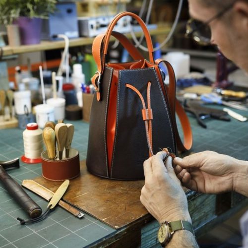 Dans son atelier, un artisan maroquinier travaille avec précision sur les finitions d'un sac seau bicolore (noir et orange). Autour de lui, l'établi regorge d'outils traditionnels comme des marteaux, des alènes et du fil de couture, témoignant d'un savoir-faire artisanal authentique. Cette scène illustre la qualité et la durabilité de la fabrication manuelle, à l'opposé de la production de masse.